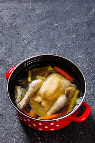 cooked chicken bouillon of whole chicken, celery, onion and carrot in red pot with ingredients and tagliatelle pasta on concrete table, vertical view, free space