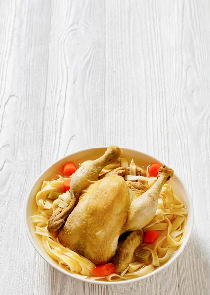 hot chicken soup of whole chicken, egg noodles, celery, onion and carrot in white bowl, vertical view, free space