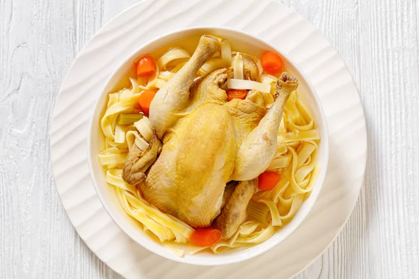 hot chicken soup of whole chicken, egg noodles, celery, onion and carrot in white bowl, flat lay, close-up