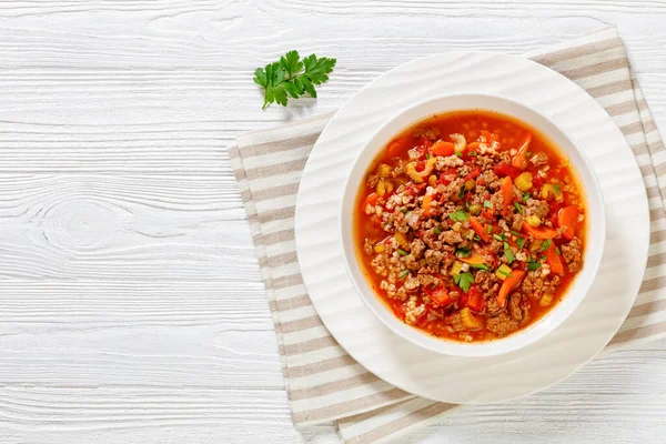 Easy hot and hearty Hamburger Soup with barley and vegetables in white bowl on white wood table, horizontal view from above, flat lay, free space