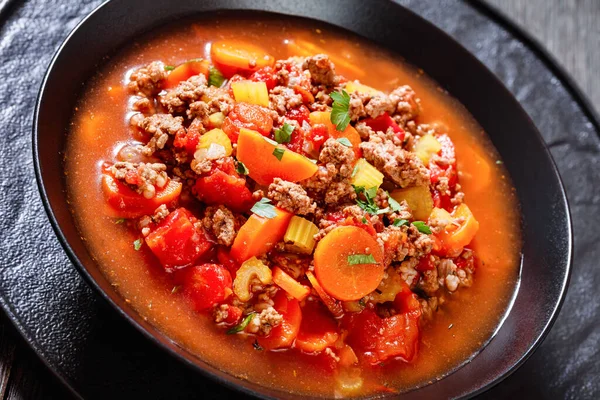 hearty Hamburger Soup with barley and vegetables in black bowl on dark wood table, horizontal view from above, fclose-up