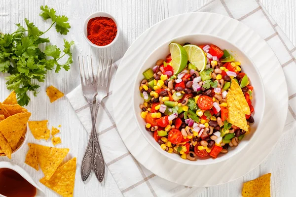 Black Bean Salad with Black-Eyed Peas, pepper, red onions, corn, cherry tomatoes and cilantro in white bowl with tortilla chips, nachos and vinaigrette dressing on wood table, landscape view, flat lay