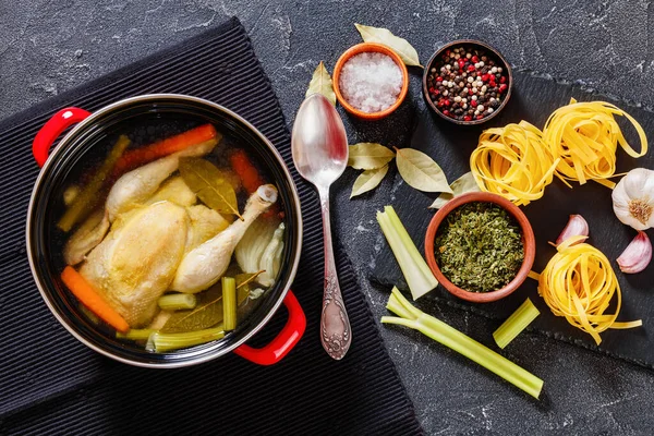 cooked chicken bouillon of whole chicken, celery, onion and carrot in red pot with ingredients and tagliatelle pasta on concrete table, landscape view from above, flat lay