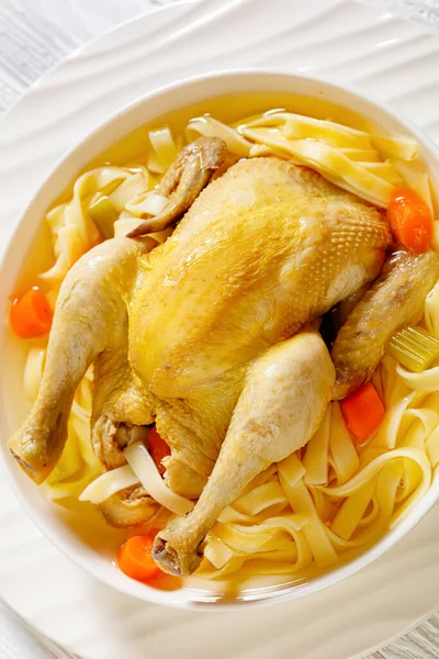 close-up of hot chicken soup of whole chicken, pasta, celery, onion and carrot in white bowl