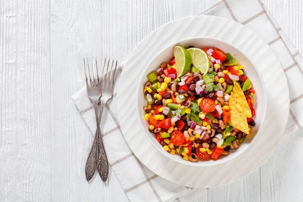 Black Bean Salad with Black-Eyed Peas, pepper, red onions, corn, cherry tomatoes and cilantro in white bowl with tortilla chips, nachos  on wood table, flat lay, free space