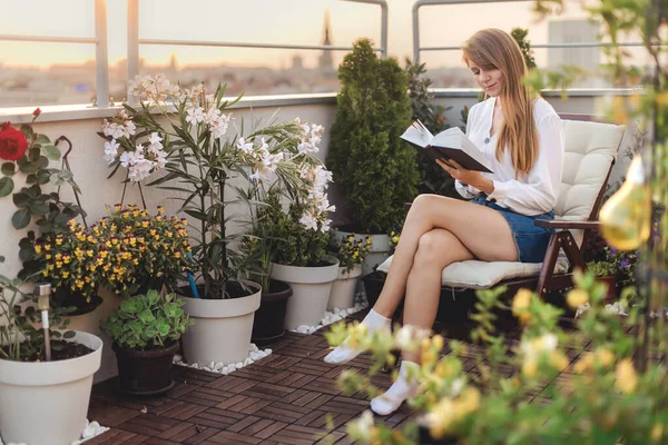 Young woman reading a book on urban rooftop garden. Girl with paper storybook sitting in chaise longue on cozy terrace with flowers, plants and city view. Female enjoying life on cozy green balcony.