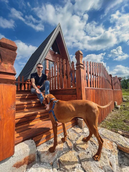 Young man sitting on front porch of wooden house with his dog. Male playing with hungarian vizsla pointer near tiny wooden house in sunny summer morning. Man traveler with pet resting in countryside.