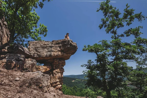 Şapkalı genç bir kadın Gospodarev kamen 'de oturuyor ve Sırbistan' da doğal cazibenin tadını çıkarıyor. Kısa şortlu bir kadın kayalık yamaçlardaki büyük kayalıklarda panoramik manzaraya bakıyor. Ormanla çevrili..