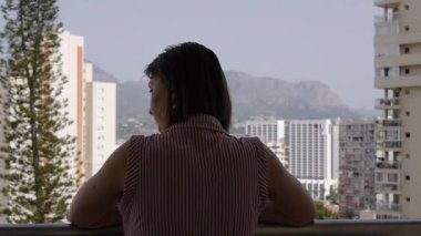 Young woman enjoying summer vacation on the balcony. Back view from the terrace of skyscrapers on mountain background.