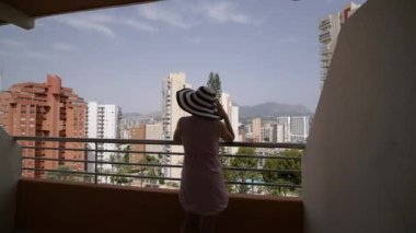 Young woman in a wide-brimmed hat enjoying summer vacation on the balcony. Wide shot of the terrace, skyscrapers and mountains.