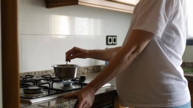 Genç hamile, beyaz tişört giyen, evdeki çelik kasede yemek karıştıran beyaz bir kadın. Mutfak sobasının yanındaki kadın karnının yakın çekimi. Yavaş çekim