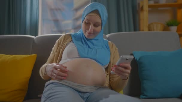 Ciężarna młoda muzułmanka w hidżabskim szaliku siedzi na kanapie za pomocą smartfona, wyszukiwarka internetowa — Wideo stockowe