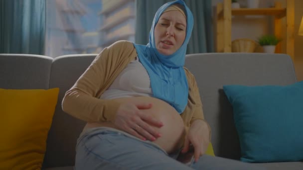 Uma mulher muçulmana grávida em cachecol está sentado no sofá e experimentando desconforto de perto — Vídeo de Stock