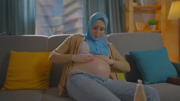 Ciężarna muzułmanin młoda kobieta używa maści lub balsam do ciała siedzi w salonie w domu — Wideo stockowe