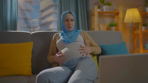 Kanepede oturan eşarplı hamile Müslüman kadın rahatsızlık ve rahatsızlık yaşıyor. — Stok video