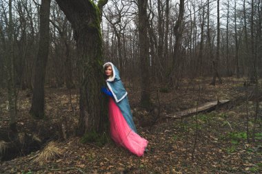 Mantolu bir kadın ağaca yaslanmış. Pelerinli fantezi kostümlü yetişkin bir kadın. sihirli sonbahar