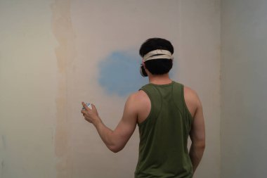 Duvara boya sıkan bir adam. Elinde sprey var. Yüzü solunum cihazında. Yenileme çalışmaları