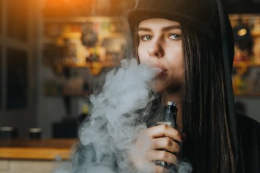 Şapkalı genç bir kadın vape dükkanında elektronik sigara içiyor. Hip-hop tarzı. Yakın çekim..