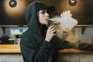 Şapkalı genç bir kadın vape dükkanında elektronik sigara içiyor. Hip-hop tarzı. Yakın çekim..