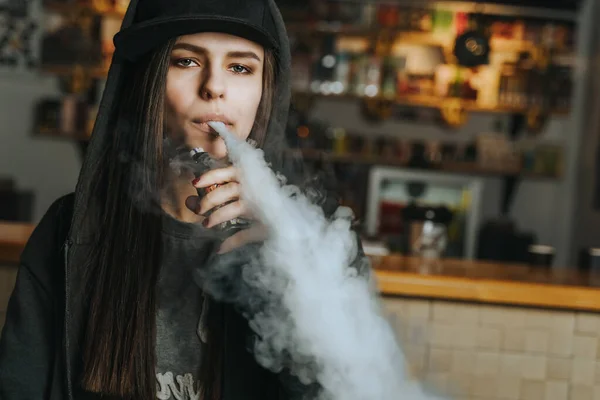 stock image Young pretty woman in cap smoke an electronic cigarette at the vape shop. Hip-hop style. Closeup.