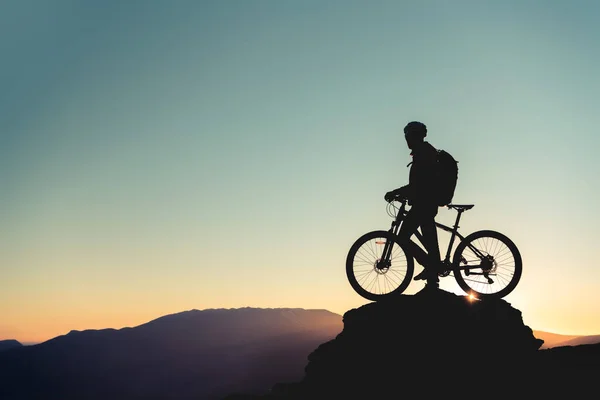 Sílhueta de ciclistas em rocha grande contra o pôr do sol Imagens Royalty-Free