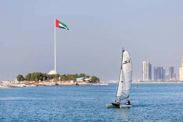 Abu Dhabi, Emirados Árabes Unidos - 5 de novembro de 2021: Dia ensolarado em Abu Dhabi Corniche com barco à vela em primeiro plano e bandeira dos Emirados Árabes Unidos ao fundo — Fotografia de Stock