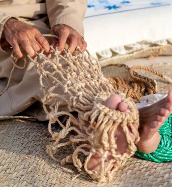 Yaşlı adam geleneksel balıkçı ağı örüyor, elleri ve ayakları çerçevede.