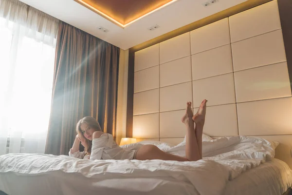 Frumoasă blondă cu păr lung într-o cămașă de bărbat își atârnă picioarele pe pat dimineața Fotografie de stoc