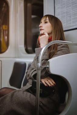 Uzun kahverengi saçlı, zarif bir ceket ve atkısı olan beyaz bir kadın metro vagonunda tek başına oturuyor ve uzaktan yan bakıyor.