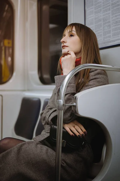 Uzun kahverengi saçlı, zarif bir ceket ve atkısı olan beyaz bir kadın metro vagonunda tek başına oturuyor ve uzaktan yan bakıyor.