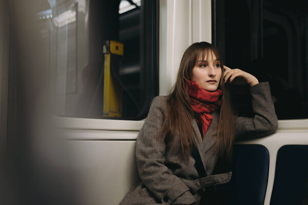 молодая кавказская женщина с длинными каштановыми волосами в элегантном стильном пальто и шарфе, сидящая одна в вагоне метро и отдаленно смотрящая в окно. Изображение с выборочной фокусировкой и тонировкой. 