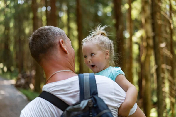 Cute baby girl making funny face on father hands.Man with backpack carry daughter in forest — Foto Stock