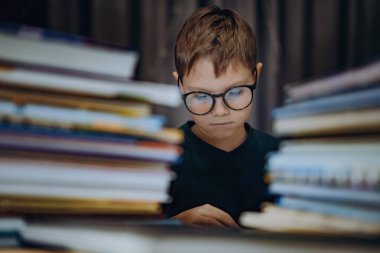 Kitap okuyan, gözlüklü, beyaz bir çocuk. Neşeli çocuk kitap yığınlarının arkasında oturuyor. Seçici odak ve gürültü efektli resim