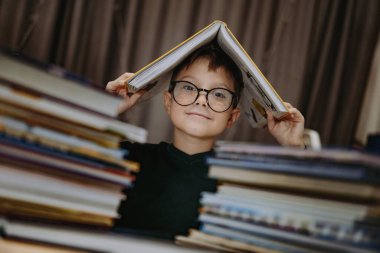 Kafkasyalı yakışıklı, gözlüklü, kafasını kitapla kaplayan bir çocuk. Kitap yığınlarının arkasından gözetleyen neşeli bir çocuk. Seçici odak ve gürültü efektli resim