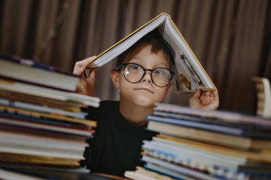 Kafkasyalı yakışıklı, gözlüklü, kafasını kitapla kaplayan bir çocuk. Kitap yığınlarının arkasından gözetleyen neşeli bir çocuk. Seçici odak ve gürültü efektli resim