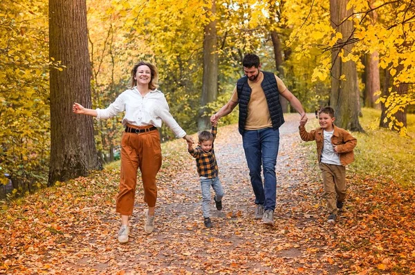 Famiglia felice: madre, padre e due figli corrono lungo il parco autunnale tenendosi per mano. — Foto Stock