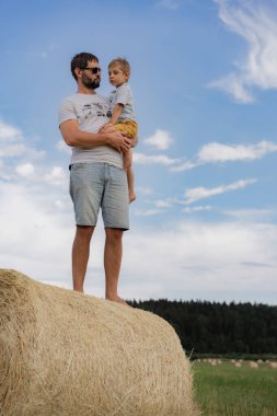 Güneşli bir yaz gününde yeşil bir tarlada yuvarlak samanlıkta oturan insan ve küçük oğlunun portresi. Yüksek kalite fotoğraf