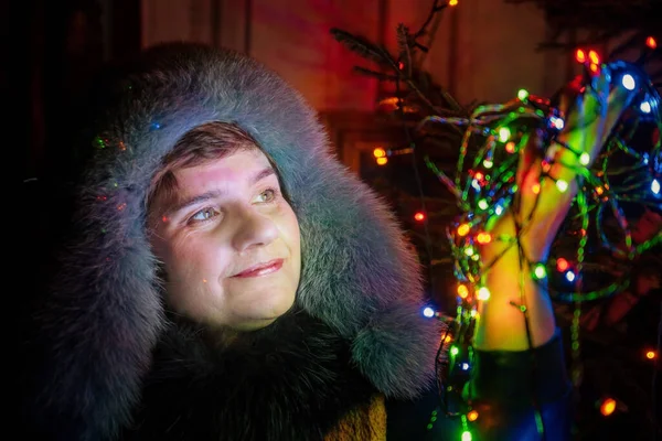 Kışın giyinmiş bir kadın Noel ağacının yanındaki renkli ışıkları elinde tutar.