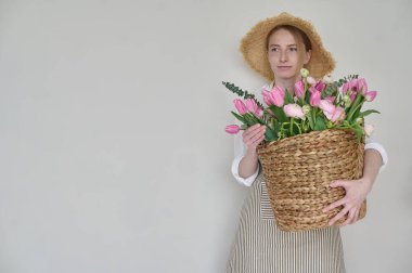 Elinde bir sepet çiçekle ve beyaz arka planda hasır bir şapkayla güzel bir çiçekçi.