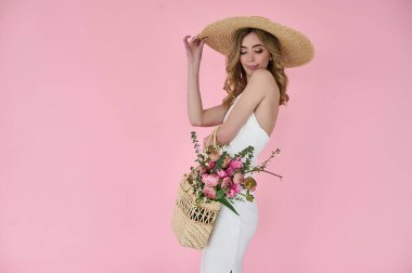 Elinde çiçek dolu hasır bir çanta ve pembe arka planda hasır bir şapkayla güzel bir genç bayan.