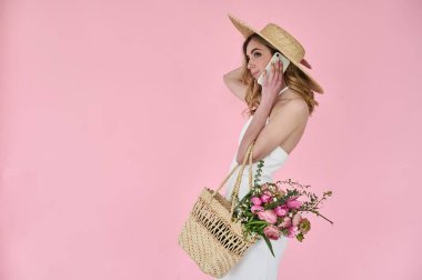Elinde çiçek dolu hasır bir çanta ve pembe arka planda telefonla konuşan hasır bir şapkayla güzel bir genç bayan.