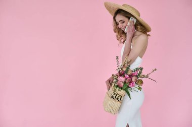 beautiful young lady with a wicker handbag of flowers in her hands and a straw hat speaking the phone on a pink background