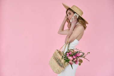 Elinde çiçek dolu hasır bir çanta ve pembe arka planda telefonla konuşan hasır bir şapkayla güzel bir genç bayan.