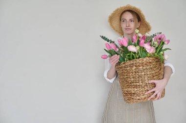 Elinde bir sepet çiçekle ve beyaz arka planda hasır bir şapkayla güzel bir çiçekçi.