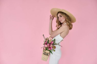 Elinde çiçek dolu hasır bir çanta ve pembe arka planda hasır bir şapkayla güzel bir genç bayan.