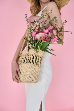 Elinde çiçek dolu hasır bir çanta ve pembe arka planda hasır bir şapkayla güzel bir genç bayan.