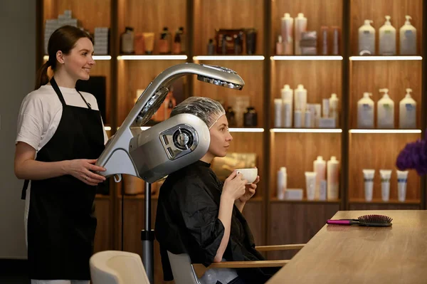 young woman under hooded dryer machine in hair salon