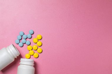 Mavi ve sarı ilaç hapları ya da vitaminler pembe arka planda beyaz kavanozlardan dökülüyor.