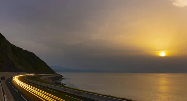 Car lights along the coast road with the sun at dusk.