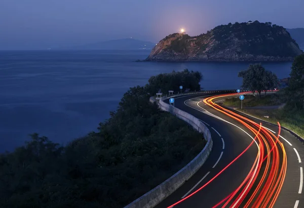 Car lights on the road on the coast of Euskadi. Getaria lighthouse at dusk.